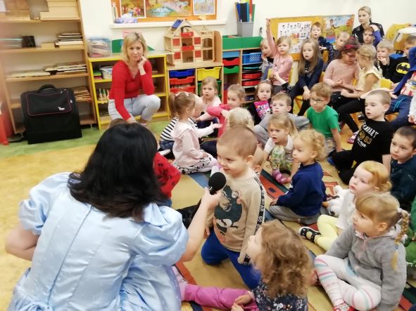Grupa dzieci z nauczycielkami w sali, z lewej strony u dołu pani w niebieskiej sukience trzyma mikrofon przed jednym z dzieci.