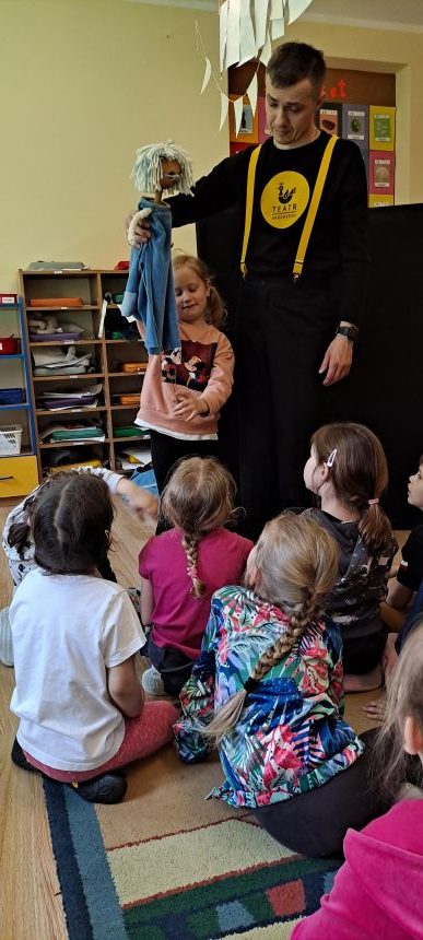 Pan w czarnym ubraniu trzyma lalkę o jasnych włosach i niebieskim stroju, obok stoi diewczynka w różowym ubraniu, grupa dzieci w sali przedszkolnej.
